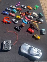 Overview of all toys including silver remote control car, various small toy cars, figures, and animals on black blanket outdoors