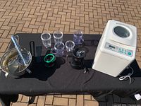 Wide view of all four items on a black cloth, showing breadmaker, blender with cups, pot, and potato masher/grinder