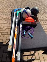 Photo showing three hockey sticks, two balls (small football and basketball), orange training cones, and a purple skipping rope on a table.