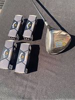 TaylorMade Burner 3 wood golf club shown with the clubhead and four unopened boxes of Callaway Warbird golf balls on a dark surface.