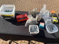 Overview photo showing multiple containers, jars, and bags filled with various nails including deck nails, roofing nails, and galvanized nails laid out on a black surface.