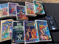 Photo showing 9 kids VHS tapes and 1 loose black VHS tape on a table, titles include Babe Pig in the City, The Sword in the Stone, Pinocchio, Snow White, Tarzan, Peter Pan, Buzz Lightyear, Lady and the Tramp, and Toy Story.