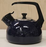 Side view of blue enameled kettle showing bulbous shape, black handle, metal lid with black knob, whistle spout with flip cap.