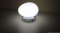Egg-shaped light fixture lit, showing the glowing white egg diffuser on a chrome round base with on/off switch.