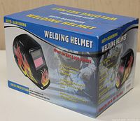 Photo of the full boxed welding helmet showing flame graphics and product features including solar powered rechargeable backup battery, full face coverage, ratcheting headband, automatic power on/off with adjustable delay time, and UV/IR protection.