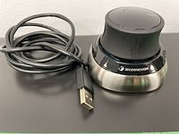 Wireless 3DConnexion SpaceMouse with USB charging cable on black surface, showing front angled view with USB cable unplugged.