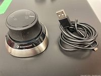 3DConnexion Space Mouse with charging cable coiled beside it, showing the device from an angle.