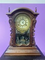 Full front view of the antique wooden mantel clock showing carved wood details, round clock face with Roman numerals, and a glass door with etched scene.
