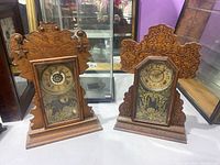 Pair of two carved wooden mantel clocks with glass fronts showing painted decorations, displayed side-by-side.