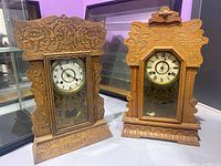Pair of antique wooden mantel clocks with intricate carvings, one with Arabic numerals and the other with Roman numerals, glass front doors open showing pendulums and keys inside