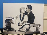 Large black and white canvas print showing a seated couple surrounded by stacks of hardcover books on either side.