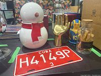 Photo showing a white ceramic snowman cookie jar with a red scarf, gold-tone metal decorative hand, a football-themed nesting doll of Kurt Warner, a glass jar with palo santo wood sticks, and a red Cuba license plate.