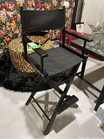 Front view of a black wooden director's chair with black fabric seat and backrest. Chair includes armrests and foot rest. Chair is positioned in a room with colorful floral wallpaper and leopard print cushion.