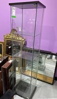 Tall glass display cabinet with veneer top and base visible from front corner angle showing open interior glass shelves and base tier.