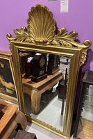 Full view of the ornate gold framed wall mirror showing the carved shell and leaf design at the top, reflecting antique clocks and furniture.