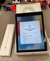 iPad 6th Generation turned on displaying country selection screen in original box, Apple Pencil box next to it.