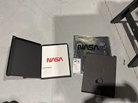 NASA Graphic Standards Manual open in a black folder, two sealed notebooks in black boxes wrapped in plastic.