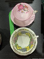 Top view of both teacups placed inside their saucers on a black surface, showing a pink teacup with floral bouquet and a white teacup with colorful floral wreath.