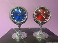 Pair of atomic style clocks by Strauss displayed side by side on a dark table with pink wall background. One clock has a blue face and one a red face, both with chrome stands.