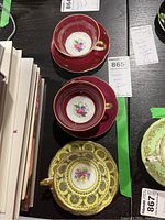 Three porcelain teacups and saucers on display, two red and one yellow decorated with black, all with floral patterns and gold trim.