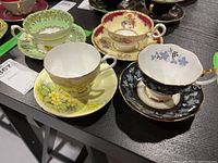 Overview of all four teacups and saucers showing varied floral designs and colors including black grapevine pattern, yellow floral, and cream with crimson rim.