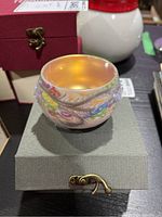 View of teacup resting on gray keepsake box, showing colorful exterior decoration and amber interior glaze.