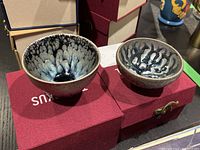Two Tenmoku glazed teacups visible inside open red Tenmokus branded keepsake boxes showing black and metallic glaze pattern interiors.