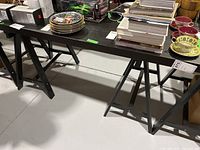 Black trestle table with rectangular top holding various books and dishware, showing condition of top and trestle legs.