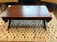 Front view of rectangular antique style coffee table with leather top embossed with decorative gold detailing, showing the central drawer with brass hardware and curved X-shaped legs with brass feet.