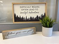 Framed sign with quote 'Difficult roads often lead to beautiful destinations', wooden block sign 'follow your own path', and artificial plant on desk