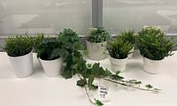 Nine artificial green plants arranged on a white table, varying in style and shape with white ceramic pots, seen from above and front angle.