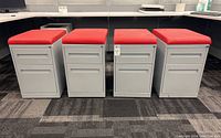 Four gray metal filing cabinet drawers on caster wheels aligned side by side, each topped with removable red vinyl cushions attached by Velcro.