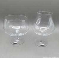 Two large clear crystal snifters with original stickers on their bases. One snifter has a wide bowl and short stem, the other a bulbous bowl and longer stem.