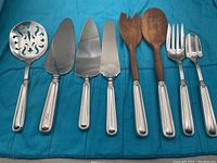 Eight serving pieces arranged on cloth: slotted scoop, three servers, two wooden salad servers, fork, scoop