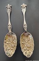 Two silver plated berry spoons showing full length, matched pair with fleur-de-lis pierced handles and grape cluster embossed bowls.