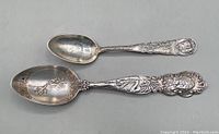 Top view of both antique souvenir spoons laid side by side showing size difference and details on handles and bowls.
