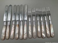 Flat lay of 6 dessert knives and 6 dessert forks with mother of pearl handles and engraved metal parts.