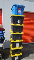 Stack of six black HDX Tough Totes with yellow lids topped by one blue Rubbermaid Roughneck bin in front of an orange roll-up door.