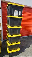 Stack of six black HDX Tough Tote bins with bright yellow lids, outdoors against storage unit door.
