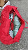 Red fabric carry bag partially unzipped, showing tree sections wrapped in plastic inside bag.