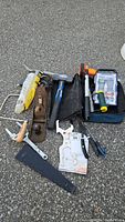 Overview of various hand tools including Dremel rotary tool kit, adjustable wrench, planer, sledgehammer, Stanley glue tool, and other tools on asphalt ground.