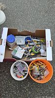 Cardboard box with mixed hardware and tools including screws, tape, scrapers, and hand tools, plus two buckets filled with assorted small hardware pieces