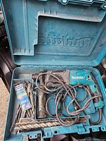 Open teal Makita carrying case showing blue rotary hammer drill, coiled cord, black side handle and bit compartment
