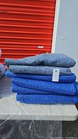 Stack of 6 moving blankets showing 4 blue quilted blankets and 2 grey wool-like pads with a '30' tag.