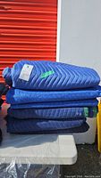 Six blue moving blankets stacked on a white surface outside a red door or shutter.