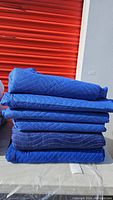 Stack of six folded blue moving blankets with visible quilting pattern and some wear.
