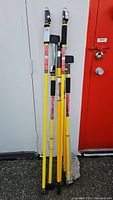 Two yellow telescoping support rods with black grips and adjustable clamps leaning against a wall next to a red door.