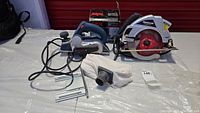 Lot of three power tools on a light surface with a red background: Mastercraft Hawkeye circular saw, Bosch electric planer PL1682, Skil palm grip electric sander in box, all cords attached.