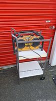 DeWalt worksite radio placed on top of metal cart in front of red storage door, showing the entire cart and radio setup.