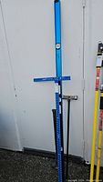 Photo of 48 inch blue T-Square, sledge hammer, and yellow magnetic sweeper leaning against a white wall.